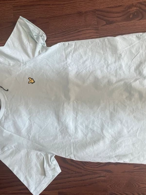 Ljus blå T-shirt lyle scott  - Snygg vit t-shirt från Lyle & Scott med en liten gul fjärilslogga på bröstet. Klassisk passform och rund halsringning. Perfekt basplagg till garderoben.