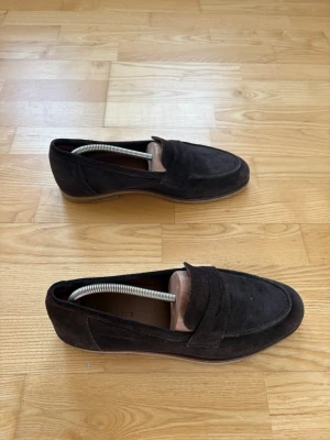 Mörkbruna pennyloafers i mocka - Snygga mörkbruna loafers i mocka med klassisk pennyloafers-design. Skorna har låg klack och en stilren siluett som passar till många outfits. Perfekta för dig som gillar en clean och tidlös look. Märke: there glory days. Nypris: 1600. Passar 42-43