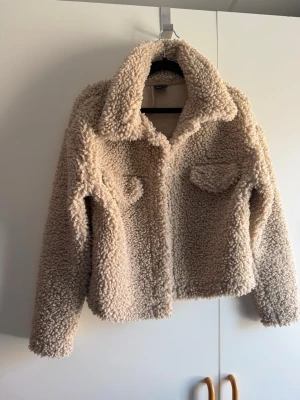 Beige teddyjacka från Gina Tricot - Supermjuk och fluffig teddyjacka i beige från Gina Tricot. Jackan har stora krage, två bröstfickor med lock och stängs med tryckknappar. Perfekt för dig som vill ha en trendig och mysig look under kyliga dagar. Köpt begagnad, katt finns i hem!