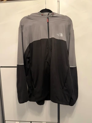 Grå och svart vindjacka The North Face - Snygg vindjacka från The North Face storlek Xl men passar L lika bra i tvåfärgad design med grå upptill och svart nedtill. Jackan har huva, dragkedja framtill och logga på bröstet. Perfekt för blåsiga dagar och enkel att matcha med olika outfits.