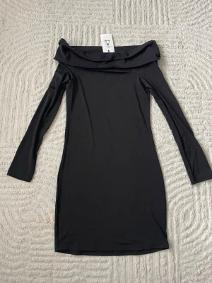 Svart offshoulder klänning - Trendig svart offshoulder klänning med lång ärm och figurnära passform. Klänningen har en bred krage som vilar över axlarna och är tillverkad i ett mjukt, stretchigt material som ger en snygg siluett.