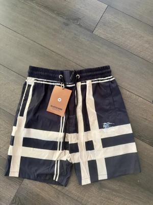 Burberry rutiga badbyxor blå/vit - Stilrena badbyxor från Burberry i marinblått och vitt med klassiskt rutigt mönster. De har elastisk midja med snörning och sidofickor. Perfekta för stranden eller poolen och ger en lyxig känsla till din sommarstil. Storleken är M men passar mer S skulle jag säga