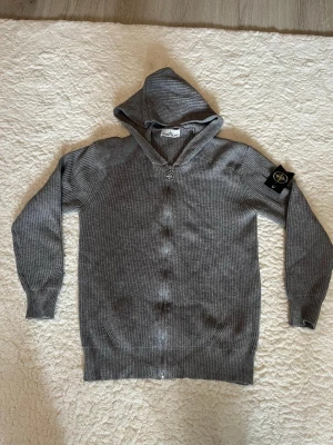 Grå hoodie från Stone Island - Säljer en grå ribbstickad hoodie från Stone Island med dragkedja framtill och ikonisk svart-gul patch på ärmen. Hoodien har långa ärmar och en skön huva. Perfekt för dig som gillar streetwear och vill ha något stilrent men ändå med edge.