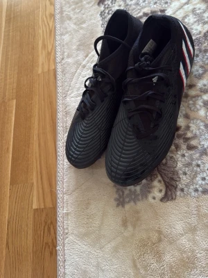 Svarta Adidas fotbollsskor med ränder(använd 1 gång) - Säljer ett par svarta Adidas fotbollsskor med klassiska vita och röda ränder på sidan. Skorna har snörning och en modern, mönstrad ovandel. Sulan är svart med blå dobbar, perfekt för spel på gräs. Snygg och sportig design för dig som vill sticka ut på planen.