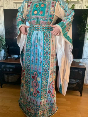 Traditionell långklänning med broderi - Unik långklänning i ljusblått och beige med färgstarka broderade mönster i rött, grönt och gult. Klänningen har vida ärmar och ett vackert bälte i midjan. Perfekt för dig som vill sticka ut med en färgglad och detaljerad look.