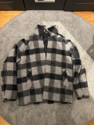 Rutig overshirt från Peak Performance - Snygg overshirt från Peak Performance i grått och svart rutmönster. Tillverkad i ett mjukt, ulligt material med två sidofickor och knappstängning vid ärmsluten. Svart innerfoder och diskret logga på ärmen ger en clean look. Helt ny aldrig använd.