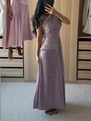 Ljuslila långklänning med drapering - Elegant långklänning från Bella Barnett i ljuslila med vackra draperade detaljer över axel och midja. Klänningen har omlottkänsla upptill och ett långt, svepande fall. Använd 1 gång. Storlek S. Nypris 1000:-  