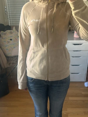 Beige velour hoodie - Säljer en beige velour hoodie från Juicy Couture med dragkedja framtill, två fickor och justerbar huva med snören. Klassisk logga broderad på bröstet och mjukt material som ger en lyxig känsla. Perfekt för en avslappnad och trendig look.