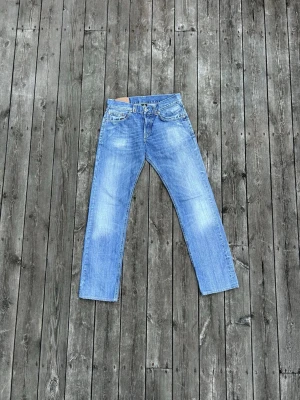 Blå raka jeans från Dondup - Säljer ett par klassiska blå jeans från Dondup med rak passform och fem fickor. Jeansen har snygga slitningar och kontrastsömmar, samt detaljer som en liten röd söm vid hällan. Perfekta för en avslappnad stil.