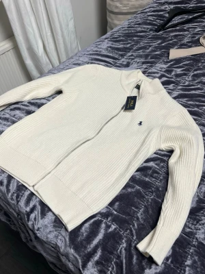 Vit stickad ziptröja Polo Ralph Lauren - Vit ribbstickad ziptröja från Polo Ralph Lauren med blå broderad logga på bröstet. Tröjan har hög krage, långa ärmar och dragkedja framtill. Perfekt för en clean och stilren look.