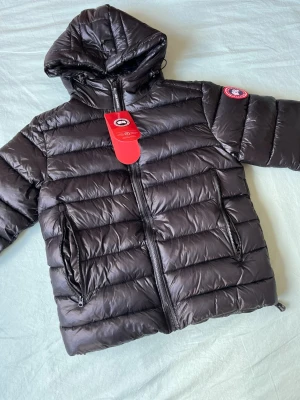 Svart dunjacka från Canada Goose - Snygg svart dunjacka från Canada Goose med huva och två dragkedjeförsedda fickor framtill. Jackan har det klassiska Canada Goose-märket på ärmen och quiltad design. Perfekt för kalla vinterdagar och riktigt varm tack vare dunfyllningen.