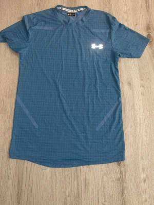 Blå träningströja från Under Armour - Snygg blå träningströja från Under Armour med diskret rutmönster och reflekterande logga på bröstet. T-shirten har rund halsringning och är tillverkad i ett lätt och snabbtorkande material, perfekt för träning och sport.
