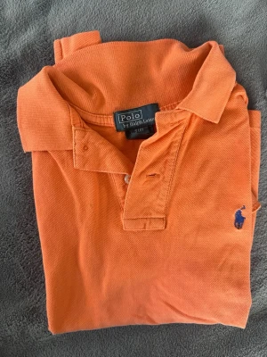 Orange pikétröja från Ralph Lauren - S i barnstorlek! Snygg orange pikétröja från Ralph Lauren med klassisk krage och knäppning framtill. Tröjan har det ikoniska blå broderade logotypen på bröstet och är tillverkad i bomull. Perfekt för en avslappnad och stilren look.
