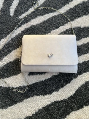 Silvrig kuvertväska med kedja - Stilren silvrig kuvertväska i sammetsliknande material med silverfärgad kedja och en sköldpadda med ”diamant” fastlimmad. Har en del sminkfläckar.