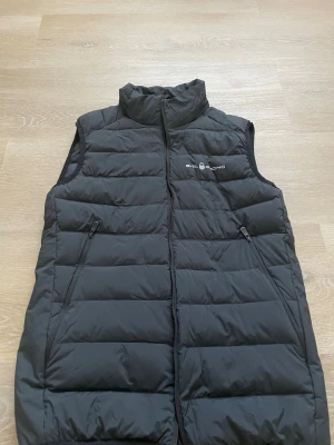 Svart dunväst från Sail Racing - Stilren svart dunväst från Sail Racing med quiltad design och hög krage. Västen har två dragkedjeförsedda fickor på sidorna och logga på bröstet. Perfekt för lager-på-lager och håller dig varm under kyliga dagar.