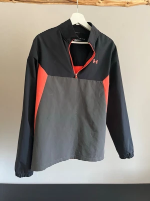 Svart och röd träningströja Under Armour - Sportig långärmad träningströja från Under Armour i svart och rött. Tröjan har halv dragkedja framtill, elastiska muddar och logga på bröstet och ryggen. Perfekt för löpning eller gym. Materialet är lätt och andas.