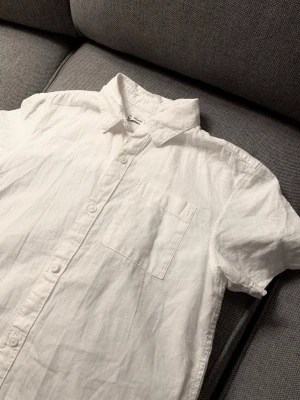 Vit kortärmad linneskjorta - Snygg vit skjorta i linne med korta ärmar och klassisk krage. Skjortan har knappar framtill och en bröstficka. Perfekt för varma dagar och enkel att matcha med olika outfits.