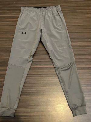Grå byxor från Under Armour - Snygga grå byxor från Under Armour.Byxorna har två sidofickor och svart logga på vänster lår. Perfekta för chill eller träning, tillverkade i mjukt material och byxorna har inte används och byxorna är S innan ni köper skriv.