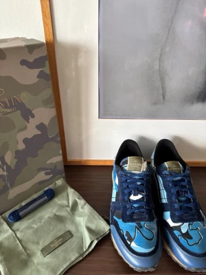 Blå camo sneakers från Valentino - Snygga sneakers från Valentino i blått camo-mönster med marinblå och ljusblå toner. Skorna har mockadetaljer, vita och svarta inslag samt gummisula. Tungan har guldfärgad logotyp och innersulan är svart. Perfekta för dig som vill sticka ut med din stil. Allt og ingår! Obs svarta prickar på hälen