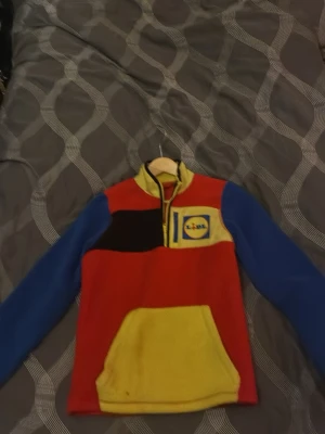 Färgglad fleece från Lidl - Fleece-tröja i rött, blått och gult med Lidl-logga på bröstet. Tröjan har halv dragkedja, blå ärmar, gul krage och magficka. Perfekt för dig som gillar färg och vill sticka ut. Mjuk och skön med sportig känsla.