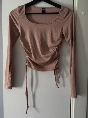 Beige ribbad topp med snörning SHEIN - Trendig beige långärmad topp från SHEIN med ribbad struktur och snörning i sidorna. Toppen har rund halsringning och croppad passform, perfekt för en snygg och avslappnad stil.