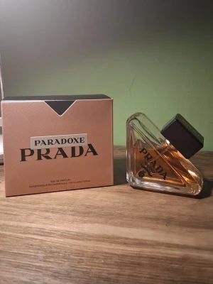 Prada Paradoxe Eau de Parfum - Prada Paradoxe Eau de Parfum med en unik trekantig glasflaska och svart fyrkantigt lock. Doften kommer i en lyxig roséguldig kartong med svart detalj. Parfymen har en elegant och modern design som sticker ut på hyllan.