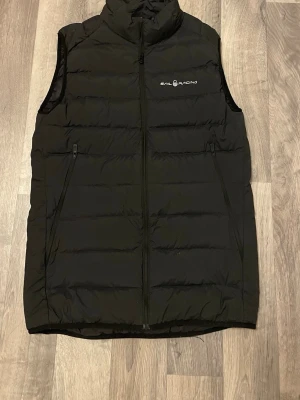 Svart dunväst från Sail Racing - Stilren svart dunväst från Sail Racing med diskret logga på bröstet. Västen har hög krage, två dragkedjeförsedda fickor och quiltad design. Perfekt för lager-på-lager och håller dig varm under kyliga dagar.