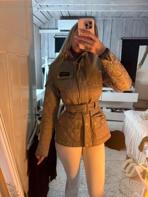 Quiltad beige jacka med bälte Barbour - Snygg quiltad beige jacka från Barbour med bälte i midjan och guldiga knappar. Jackan har lång ärm, flera fickor och klassisk Barbour-logga på bröstet. Perfekt för dig som gillar en stilren och tidlös look.