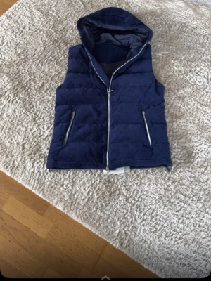 Mörkblå Manchester vest med luva - Snygg mörkblå dunväst med huva och dragkedja framtill. Västen har två fickor med dragkedja och quiltad design. Perfekt att slänga över en hoodie för en cool och avslappnad look.