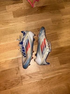 Adidas X Speedflow blå/vita fotbollsskor - Säljer ett par Adidas X Speedflow fotbollsskor i blått, vitt och rött. Skorna har snörning, låg profil och FG-dobbar för spel på naturgräs. Lätta och smidiga med syntetiskt material för maxad fart på planen. Perfekta för dig som vill sticka ut med stil och speed. 