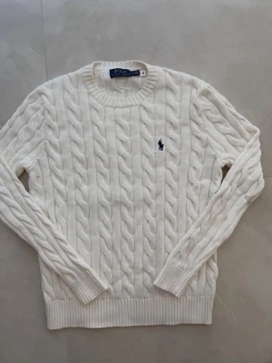 Vit kabelstickad tröja Polo Ralph Lauren - Kabelstickad vit tröja från Polo Ralph Lauren med rund halsringning och broderad marinblå logotyp på bröstet. Tröjan har långa ärmar och ribbstickade muddar samt nederkant. Perfekt för en klassisk och stilren look.