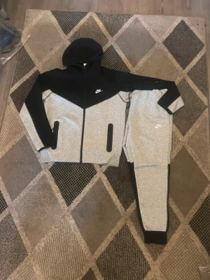 Nike Tech Fleece grå och svart set - Nike Tech Fleece set med hoodie och byxor i grått och svart. Jackan har dragkedja, huva och svarta detaljer vid fickorna. Byxorna har muddar vid benslut och vit Nike-logga. Perfekt för chill eller träning.