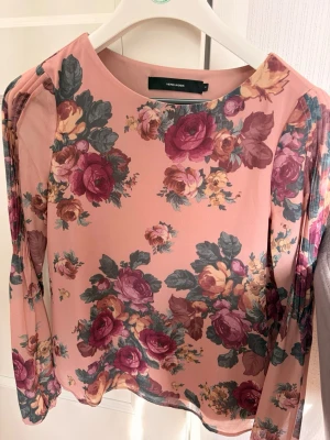 Blommig långärmad blus Vero Moda - Snygg blus från Vero Moda i ljusrosa med stort blommönster i vinrött, gult och grönt. Blusen har rund halsringning och långa ärmar med veckade detaljer vid axlarna. Perfekt för dig som gillar romantisk och färgstark stil.