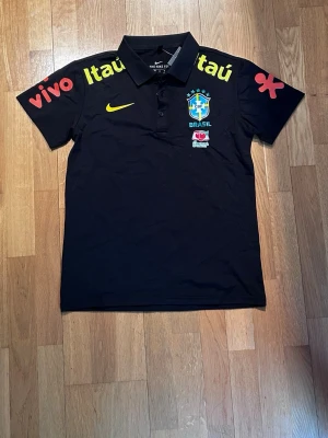 Svart Brasil Nike pikétröja - Snygg svart pikétröja från Nike med Brasilien ligga på bröstet och flera sponsortryck i gult och rött. Tröjan har klassisk krage med knappar och är perfekt för dig som gillar fotboll. Säljer då den är för liten (storlek s) och är själv storlek M!