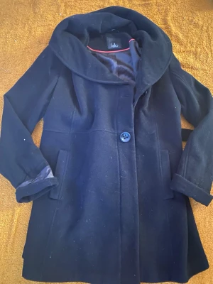 Svart kappa med stor krage från Ichi - Stilren svart kappa från Ichi med stor, mjuk krage och enkel knäppning framtill. Kappan har två fickor och är lätt utsvängd i modellen. Perfekt för kyliga dagar och ger en snygg siluett. Finns en defekt vid knappen me går lätt och fixa 