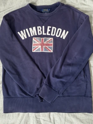 Ralph lauren sweater - Blå sweatshirt från Polo Ralph Lauren med trycket 'Wimbledon' i vitt och en brittisk flagga på bröstet. Används aldrig, tveka inte o fråga vad som om ni har några funderingar