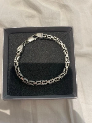 Silverfärgat kedjearmband i box - Snyggt armband i silverfärgad metall med flätad kedjedesign och karbinlås. Armbandet har ett stilrent och robust utseende som passar perfekt till en modern look. Levereras i en svart presentask.