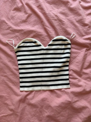 Randig bandeau topp från H&M - Snygg bandeau topp med svarta och vita ränder och tunna axelband. Toppen har en hjärtformad urringning och är croppad, perfekt för varma dagar eller sommarens outfits.