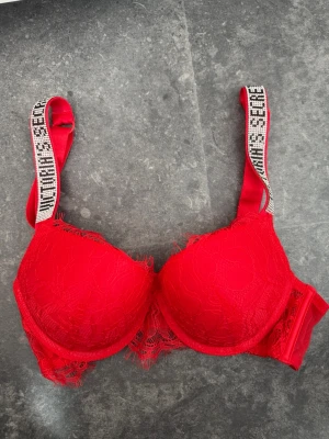 Röd spets-bh från Victoria's Secret - Snygg röd bh från Victoria's Secret med vadderade kupor och spetsdetaljer. Axelbanden är dekorerade med text och ger en sportig touch. Perfekt för dig som vill ha något extra i underklädeslådan. NYSKICK och aldrig använd. Den har pushup och är i storlek 75B!!