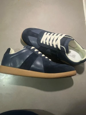 Maison Margiela sneakers blå kvitto finns  - Maison Margiela sneakers i marinblått med kontrasterande vita snören och beige gummisula. Skorna har en mix av skinn och mocka på ovandelen, rund tå och klassisk sneakerform. Snygg retrostil med diskreta detaljer och bekväm innersula.