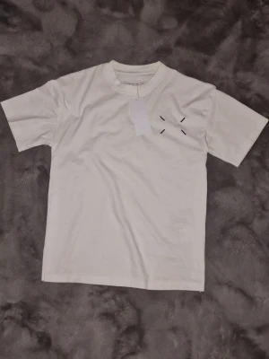 Vit Maison Margiela t-shirt - Vit t-shirt från Maison Margiela med svart tryck på ryggen och diskret broderad detalj på bröstet. Klassisk passform med rund hals och korta ärmar. Tillverkad i mjuk bomull, perfekt för en clean och stilren look. 500kr först till kvarn
