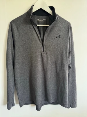 Grå halvzip tröja från Under Armour - Snygg grå långärmad tröja från Under Armour med halv dragkedja framtill och diskret logga på bröstet. Tillverkad i ett mjukt och stretchigt funktionsmaterial som passar perfekt till både träning och chill. Enkel och stilren design.