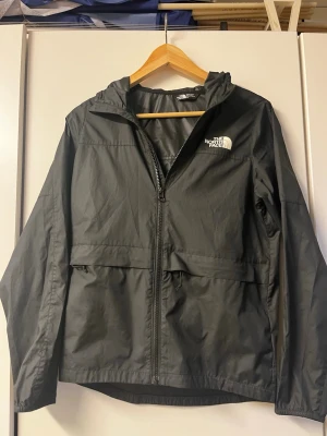 The North Face vindjacka  - Säljer en svart vindjacka från The North Face med huva och dragkedja framtill. Jackan har logga på bröstet och ryggen, två stora fickor och är tillverkad i lätt, vindtätt material. Jackan är i ett extremt bra skick då den knappt används. Säljer för 250 kr vilket är ett väldigt bra pris med tanke på skicket på jackan.