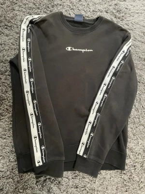 Svart Champion sweatshirt med logga - Svart sweatshirt från Champion med vit broderad logga på bröstet och vita band med Champion-tryck längs ärmarna. Klassisk rund halsringning och ribbade muddar. Perfekt för en sportig och avslappnad stil.