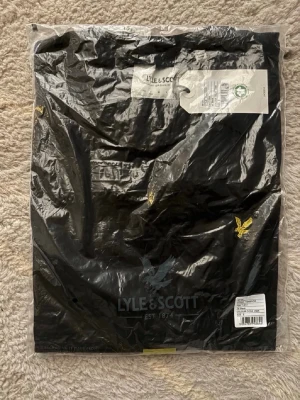 Svart t-shirt från Lyle & Scott - Svart t-shirt från Lyle & Scott med klassisk gul logga på bröstet. Tillverkad i ekologisk bomull och har en enkel, stilren design med rund hals. Perfekt för en clean och snygg look.