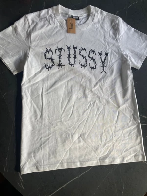Vit Stussy t-shirt med tryck - Strlk M - Vit t-shirt från Stussy med svart grafiskt tryck på bröstet. Klassisk rund halsringning och korta ärmar. Tillverkad i mjuk bomull, perfekt för en avslappnad streetwear-look.