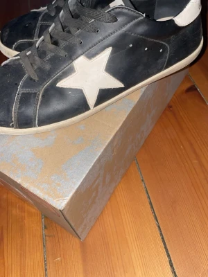 Golden Goose sneakers - Svarta sneakers från Golden Goose med vit stjärndetalj på sidan och vit häl. Skorna har svarta skosnören, vit sula och är tillverkade i läder. Klassisk låg modell med en cool och avslappnad vibe. högra skon har ett litet hål men annars är det bra