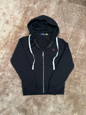 Svart hoodie från Polo Ralph Lauren S - Svart hoodie med dragkedja från Polo Ralph Lauren. Klassisk design med vit dragsko i huvan och röd broderad logga på bröstet. Mjuk insida och ribbade muddar. Perfekt för en chill och stilren look. Står M men Sitter som en S. Pris kan diskuteras 😊