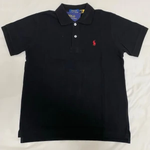 Svart pikétröja från Polo Ralph Lauren - Klassisk svart pikétröja från Polo Ralph Lauren med korta ärmar och krage. Tröjan har två knappar framtill och den ikoniska röda logotypen broderad på bröstet. Perfekt till jeans eller shorts för en clean look.