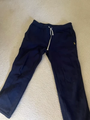 Mörkblå mjukisbyxor från Polo Ralph Lauren - Mörkblå mjukisbyxor från Polo Ralph Lauren med klassisk gul logga broderad på vänster ben. Byxorna har dragsko i midjan och är tillverkade i mjukt bomullsmaterial. Perfekta för en avslappnad stil.